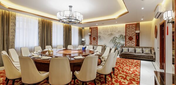 Ресторан J.Z.Peking Duck (J.Z. Утка по-пекински) . Просторная комната с круглым столом и белыми стульями, красный ковер с золотым узором, светлый потолок с подвесной люстрой, большие окна с занавесками, бежевый диван, китайская настенная роспись с текстом, декоративные панели по бокам.