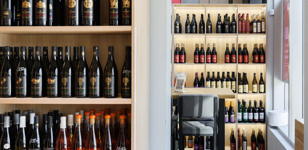 AVDRINKS (АВДРИНКС). Стеллажи с бутылками вина на деревянных полках, высокие стулья рядом с барной стойкой, современное освещение, светлые стены, витрина справа.