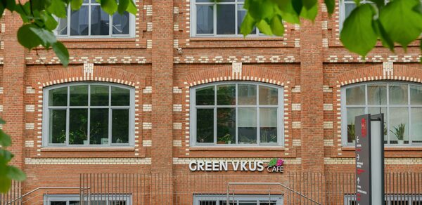 Green Loft(Зеленый Лофт). Фасад кирпичного здания с большими окнами в белой рамке. На уровне первого этажа вывеска "GREEN VKUS CAFE". Перед зданием находится металлическое ограждение и маленькая клумба. На переднем плане видны листья деревьев.