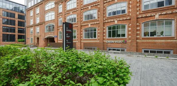 Green Loft(Зеленый Лофт). Красное кирпичное здание с большими окнами. Перед ним тротуар из серых плит и зеленые кусты. У входа вертикальная черная вывеска с текстом. На первом этаже видна надпись «GREEN VKUS».