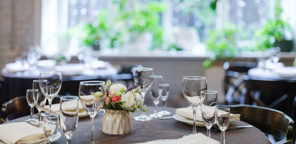 Green Loft (Зеленый Лофт). Круглый стол, сервированный белыми тарелками, стеклянными бокалами и салфетками. На столе цветочная композиция. Вокруг стулья темного цвета. На заднем плане большое окно с зелеными растениями. Естественное освещение.