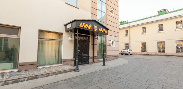 Давай Лама. Вход в здание с аркой, стеклянные окна, двери, вывеска с надписью на незнакомом алфавите.  Мощеная площадка перед входом в светлых тонах, подъездная дорожка. Фасад кремового и коричневого цветов с большим окном.