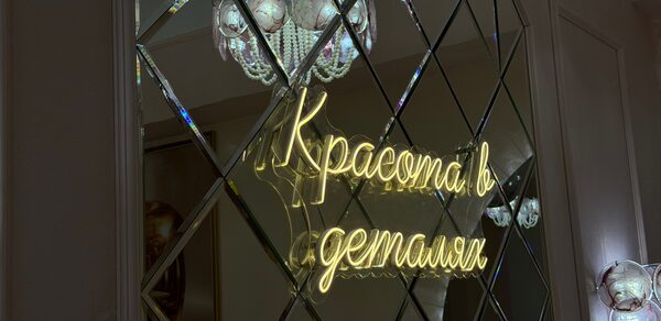Лофт «Голд Эпл». Зеркальная стена с ромбовидными панелями, неоновая надпись "Красота в деталях". Люстра с стеклянными элементами. Светлые стены, декоративное освещение.