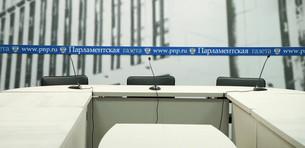Медиацентр Парламентской газеты. Конференц-зал с белыми столами, тремя черными стульями и микрофонами. На стене вывеска с текстом: "Парламентская газета, www.pnp.ru". Стены украшены серыми изображениями зданий.