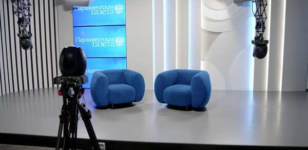 Конференц-зал до 60 гостей площадью 150 м². Студийное пространство с двумя синими креслами на сером полу, телевизионной камерой на треноге и ярким освещением. Стены оформлены белыми панелями с вертикальными полосами, присутствуют экраны с текстом «Парламентская газета».