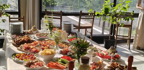 СемьСюр. Пространство с кухней до 30 гостей. Просторное помещение с панорамными окнами, стол с разнообразными блюдами, закусками и напитками. Светлое освещение, деревянные стулья, зелёные растения в горшках. На столе белая скатерть, стеклянные бокалы, тарелки. На заднем плане крупные окна с видом на зелень.