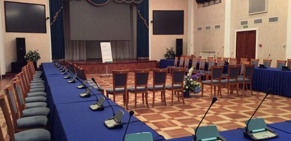 Концертный зал Ассамблея. Просторный зал с деревянным паркетным полом, столы и стулья с синими скатертями и обивкой, множество микрофонов на столах. На сцене экран с занавесом, рядом флипчарт. Освещение представлено люстрой и точечными светильниками.
