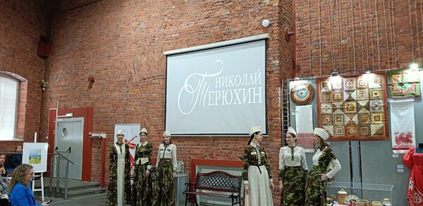 Выставочный зал Форум. Кирпичное помещение с высоким потолком. На стене проекционный экран с надписью. Слева металлические двери, справа экспонаты и яркие ткани. Несколько людей в традиционных костюмах. В зале стулья для зрителей.