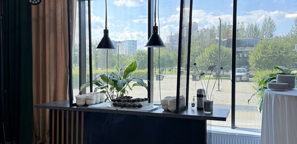 Event  Hall ARCOBALENO (Эвент Холл АРКОБАЛЕНО) . Современный интерьер с барной стойкой и подвесными черными светильниками у панорамного окна, металлическая конструкция с растениями, стеклянные стены, бежевые шторы, белая скатерть на круглом столе с тарелками.