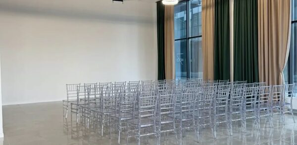 Event  Hall ARCOBALENO (Эвент Холл АРКОБАЛЕНО) . Зал с высокими потолками и большими окнами, занавешенными зелеными и бежевыми шторами. Ряды прозрачных стульев расставлены на сером плиточном полу. Две круглые подвесные лампы на черном потолке. Белая стена без украшений.