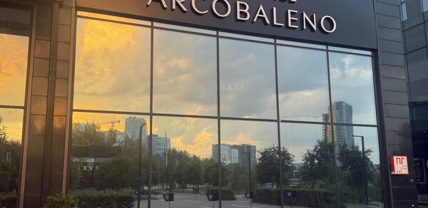Event  Hall ARCOBALENO (Эвент Холл АРКОБАЛЕНО) . Фасад здания с большими стеклянными окнами, отражающими городской пейзаж с деревьями и небоскребами. Название на фасаде: «EVENT HALL ARCOBALENO». Вывеска «П» на стекле. Цвета: черный, стеклянные поверхности отражают закатное небо.