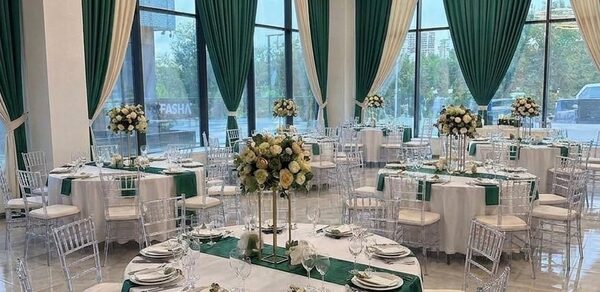 Event  Hall ARCOBALENO (Эвент Холл АРКОБАЛЕНО) . Банкетный зал с круглыми столами, украшенными белыми скатертями и зелеными дорожками. Прозрачные стулья, большие окна с зелеными шторами. Центральные композиции из светлых цветов. Потолочное освещение в виде кольцевых светильников.