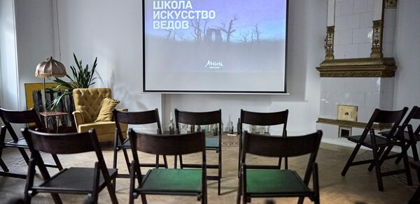 Артиста Лекторий. Комната для мероприятий с экраном, на котором проецируется текст. Стулья расставлены в ряды. В углу зелёные растения и жёлтое кресло. Светлая стена с белой плиткой и декоративной рамой. Лампа с абажуром на столике.