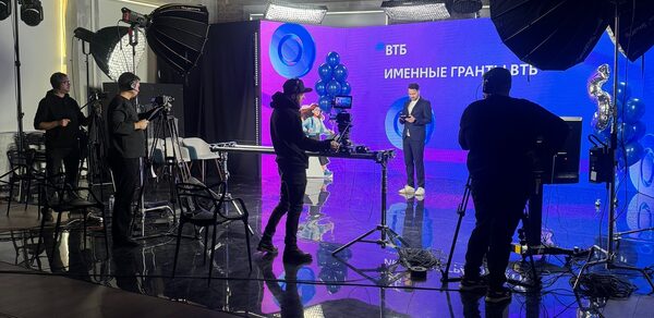 Конференц-зал до 150 гостей площадью 313 м². Студийное пространство с профессиональным освещением и видеооборудованием. Темный пол, яркий светодиодный экран с надписями, синие воздушные шары. Несколько человек работают с камерами. Стены серые, потолок с деревянными балками и светильниками.