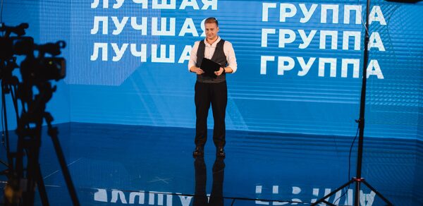 Конференц-зал до 150 гостей площадью 313 м². Мужчина в костюме стоит на сцене с синей светодиодной стеной, на которой написано "ЛУЧШАЯ ГРУППА". Черная стойка микрофона и отражения на полу. Профессиональное освещение и оборудование для съемки. Стены из светодиодов, пол глянцевый.
