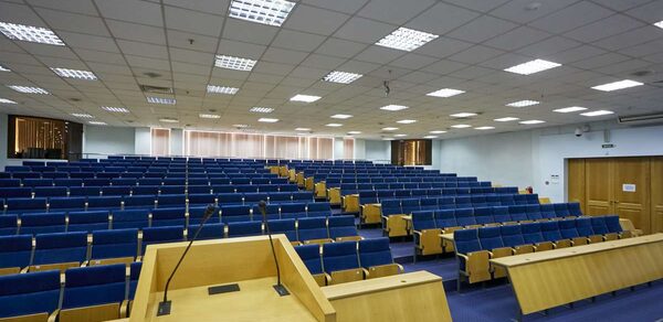 Конференц-зал Петровский. Аудиторное пространство с рядами синих кресел, деревянная кафедра с микрофонами на переднем плане. Потолочные светильники в виде квадратных решеток, стены светло-серые. У дверей на правой стороне оформлены указатели.