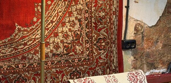 Бар Кофейно-бильярдный площадью 75 м². Настенное пространство с ковром красно-золотистого оттенка и восточным орнаментом, рядом деревянная часть стены с облупившейся штукатуркой. На переднем плане видна часть кресла с подушкой с узором. Освещение от торшера.