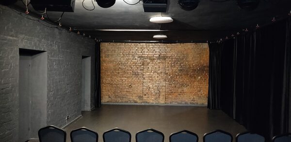 Концертный зал до 40 гостей площадью 60 м². Небольшое помещение с кирпичной стеной и серыми боковыми стенами. Потолок тёмного цвета с подвесными светильниками и прожекторами. Черные шторы по бокам. Стоят стулья рядами, темное ковровое покрытие.
