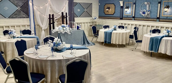Банкетный зал Сокол. Банкетный зал с круглыми столами, покрытыми белыми скатертями. Черные стулья с мягкой спинкой, декор синими и белыми элементами. Голубые и белые драпировки на стенах, круглые потолочные светильники, светлое деревянное покрытие пола.