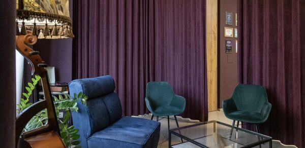 У кота до 30 гостей площадью 60 м². Гостиная с фиолетовыми шторами и тёмно-синим мягким диваном. Два кресла из зелёного материала. Стеклянный журнальный столик с чёрным металлическим каркасом. На полу плитка светлого цвета. Стены фиолетовые. Видна часть струнного музыкального инструмента и зелёное растение.