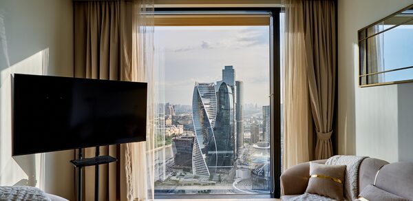 Квартира площадью 60 м² для мероприятий Свидания в Москва Сити. Гостиная с большим окном и видом на город, телевизор на стойке, светло-коричневый диван с подушками, кофейный столик, бежевые шторы и тюль, светлые стены, деревянный пол.