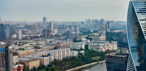 Квартира площадью 60 м² для мероприятий Свидания в Москва Сити. Вид на городскую панораму с высоты, река с лодкой, многоэтажные жилые здания разного дизайна, открытые зелёные пространства, современное стеклянное здание справа, светлое небо.