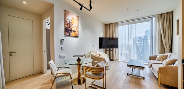 Квартира площадью 60 м² для мероприятий Свидания в Москва Сити. Современная гостиная с деревянным полом, большими окнами и светлыми шторами. Мебель включает два дивана, стол со стеклянной столешницей и стулья. На стене картина, на потолке чёрные светильники. Есть телевизор на стойке.