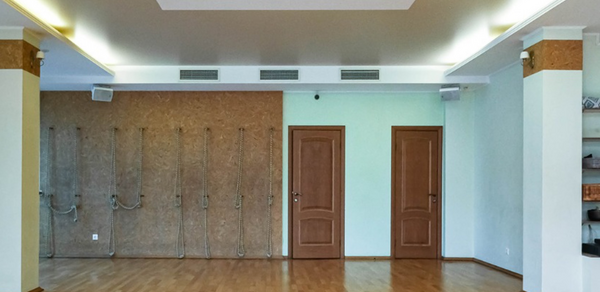 Спортзал Йога-студия площадью 100 м². Просторное помещение с деревянным полом, светлыми стенами и натяжным потолком. На одной из стен канаты и две двери. Система освещения встроена в потолок. На другой стене открытые стеллажи. Вентиляционные решетки под потолком.