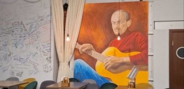 Гнездо Глухаря. Кафе с деревянными столами и разноцветными стульями. На стене большое изображение человека с гитарой. Стена покрыта светлыми шторами и надписями. Подвесные светильники создают мягкое освещение. Стена и дверь коричневого цвета.