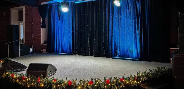 Гнездо Глухаря. Сцена с черной занавеской, освещенная прожекторами. На переднем плане новогодние гирлянды с зелеными ветвями и красными украшениями. Стены из кирпича, потолок с видимыми балками. Звуковая аппаратура по краям сцены.