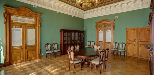 Особняк Сухозанета. Просторная комната с высокими потолками и паркетным полом, стены окрашены в зеленый цвет. Деревянные двери и мебель с классическими элементами. Пара стульев вдоль стен, круглый стол с мягкими стульями в центре. Люстра и обильный декор на потолке.
