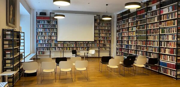 Зал для вашего мероприятия. Библиотека с деревянным полом и книжными полками вдоль стен. В центре установлены ряды стульев перед проектором и экраном. Освещение из подвесных ламп. На передней стене большие окна, справа — кресла и небольшой стол.