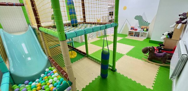 Зал площадью  122 м 2. Игровая комната с мягкими модулями и сетками, яркий пластиковый батут и горка, сухой бассейн с шариками. Напольное покрытие зелёное и бежевое, мягкие игрушки сбоку. Стены светлого цвета с рисунком динозавра и полок.