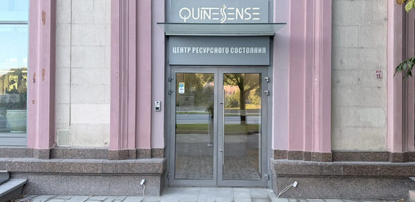 QuintesSense. Входное пространство с двумя стеклянными дверями в серых рамах. Светло-розовые и серые стены здания с текстовыми вывесками над входом. Большое окно с левой стороны. Плиточная тротуарная дорожка перед зданием.