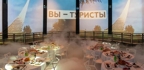 Атракцион до 80 человек. Круглое помещение с экранами, на которых изображены белые шпили и деревья на фоне синего неба. На экранах текст «ВЫ – ТУРИСТЫ». В центре стоят круглые столы с белыми скатертями, накрытые посудой и едой. Белые стулья, искусственный туман на полу.