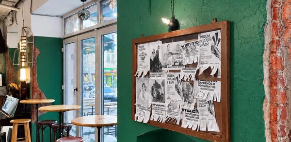 Караоке-рюмочная Шотский. Барное пространство с деревянными столами и стульями, зеленые стены с элементами открытой кирпичной кладки. На стене висит доска с печатными объявлениями. Большие окна с видом на улицу. Подвесные светильники.