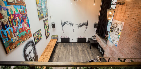 Street Art Loft (Стрит Арт Лофт). Лофт-пространство с высокими белыми стенами и кирпичной акцентной стеной. На стенах висят различные картины. Пол деревянный, из темных досок. Светильники подвешены к потолку. Помещение декорировано зеленью на ограждении.