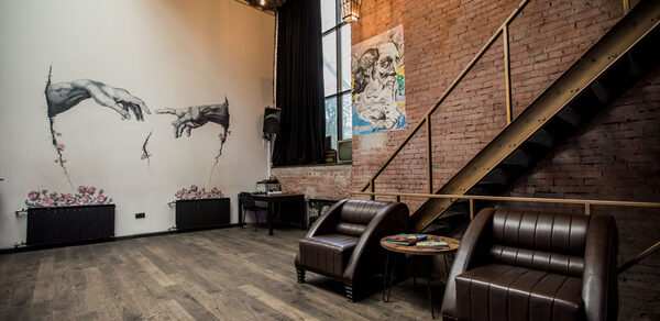 Street Art Loft (Стрит Арт Лофт). Лофт-пространство с кирпичными стенами, деревянными полами, лестницей и высокими потолками. На светлой стене граффити с пятью руками и розовыми цветами. Два кожаных кресла и круглый столик. Большое окно за закрытой шторой.