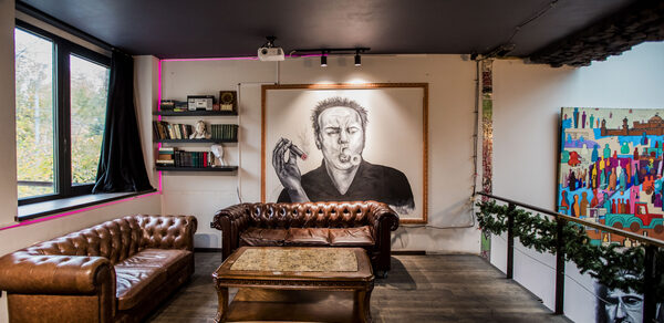 Street Art Loft (Стрит Арт Лофт). Лофт-пространство с деревянным полом, черными потолками, большими окнами, двумя коричневыми кожаными диванами, деревянным столом, настенной живописью, книжными полками и произведением искусства с изображением курящего человека.