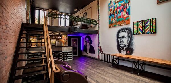 Street Art Loft (Стрит Арт Лофт). Лофт-пространство с высоким потолком, металлической лестницей и балконом. Стены украшены граффити и картинами. На полу темный ламинат. Коричневый диван у стены. Слева кирпичная кладка, справа белая стена с цветными картинами.