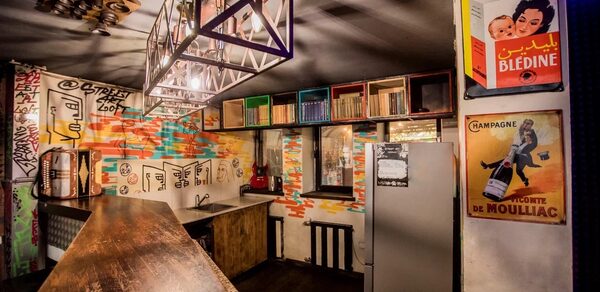 Street Art Loft (Стрит Арт Лофт). Барное пространство с деревянной стойкой, стеллажами с книгами и плакатами на стенах. Потолочные светильники в металлической раме. Стены с граффити и яркими акцентами. Холодильник серого цвета. Тёмный пол, подчеркнутый освещением.