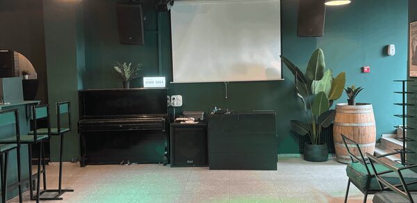 Work Shop Loft(Воркшоп Лофт). Зал 11 . Пространство с темно-зелеными стенами, проектором и экраном, задними колонками. Черное пианино, барные стулья и столы, декор в виде растений в горшках и деревянной бочки. Кафельный пол, подвесные светильники.