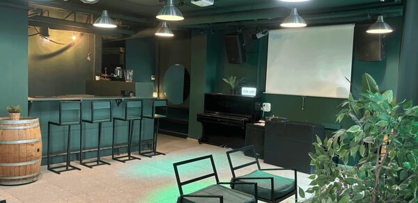 Work Shop Loft(Воркшоп Лофт). Зал 11 . Открытое помещение с барной стойкой и высокими стульями, фортепиано, экран для проектора, металлические подвесные светильники, деревянная бочка, черные стулья с мягкими подушками, зеленые стены и потолок, комнатное растение, бежевый пол.