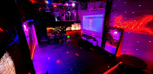 Soul Loft на Бауманской. Тускло освещенное пространство с разноцветными огнями, баром и большим экраном. На стене написано слово Soul в ярко-красных огнях, что создает живую атмосферу, подходящую для встреч или мероприятий. Деревянный пол добавляет теплоты современному декору.