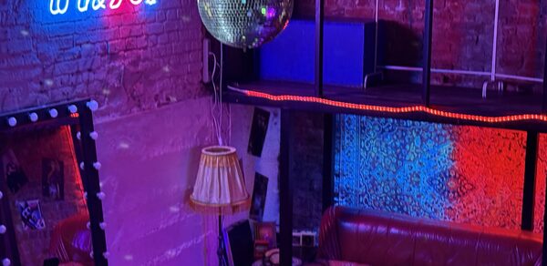 Лофт ИЗНАНКА. Зал ЛИХИЕ 90-е. Затемнённая комната лофта, много людей в кадре проводят фуршетное мероприятие, беря еду с объединённых столов. Несколько людей на кадре танцуют