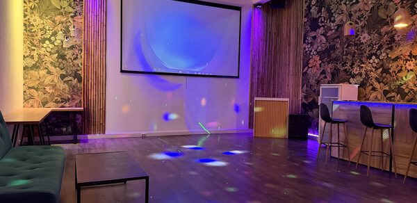 Лофт для вечеринок с коктейльным баром . Пространство с деревянным полом, стенами с цветочным узором и крупным проектором. Барная стойка с высокими стульями, справа. Диван и стол слева. Разноцветное освещение и световые проекции на полу.