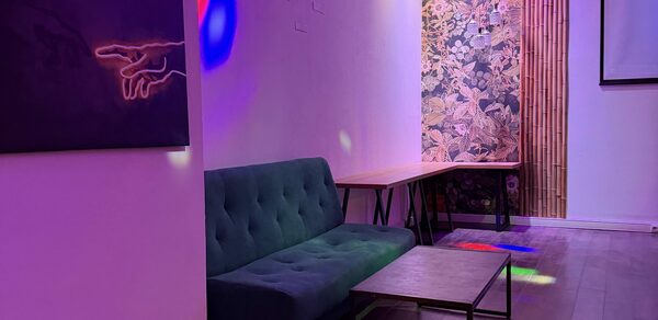 Лофт для вечеринок с коктейльным баром . Гостиная с зелёным диваном, деревянным столом и кофейным столиком. Стена украшена яркими обоями с растительным узором и рядом бамбуковых панелей. Висящая картина показывает руку. Цветное освещение, кирпичная и штукатуренная стены. Паркетный пол.