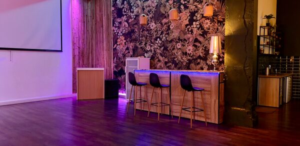 Bar on Presnya for private events. Просторное помещение с деревянным полом, на стене большой экран. Справа барная стойка с тремя стульями, зона украшена обоями с цветочным узором и подвесными светильниками. Левую часть зала освещают фиолетовые и синие огни.