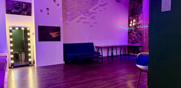 Bar on Presnya for private events. Просторное помещение с деревянным полом, фиолетовым освещением, белыми и черными стенами с графикой. Присутствуют синий диван, столы, барный стул и зеркало с подсветкой. На потолке светильник в виде люстры.