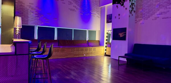 Bar on Presnya for private events. Просторное помещение с деревянным полом, фиолетовой подсветкой, барной стойкой и тремя стульями. У стены диван, возле кирпичной колонны — лампа на стойке. Стены декорированы белой кирпичной кладкой, на потолке подвесная люстра.